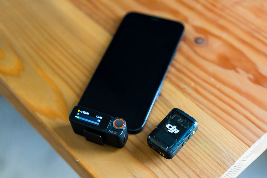 A smartphone, a small black electronic device, and a compact rectangular gadget with a screen are placed on a wooden surface.