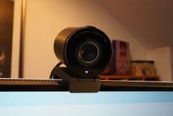 A black webcam mounted on top of a computer monitor, with a visible lens and a small light indicator below it, against a blurred background.