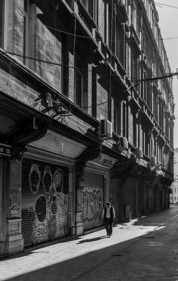 A person walks alone along a narrow, deserted street lined with shuttered, graffiti-covered shops in a black-and-white urban scene.