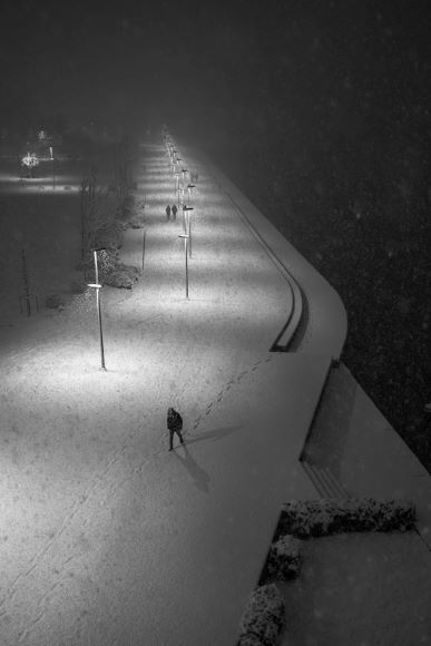 A solitary figure walks on a snow-covered path lined with streetlights at night. Other distant figures are visible further along the path under the illuminated snow.