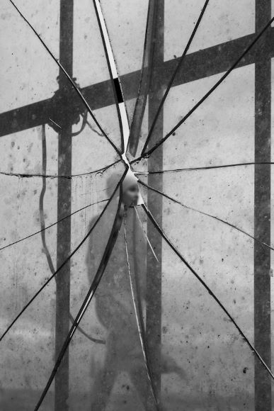 Cracked glass panel with an indistinct figure partially visible through the center, against a backdrop of intersecting lines and shadows.