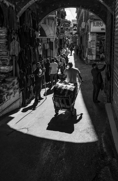 A person pushes a cart through a narrow, sunlit street lined with shops and clothes displays, under an arched entrance. Shadows and pedestrians are visible. Black and white image.