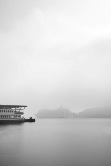 Foggy waterfront scene with a boat docked on the left. Two people stand at the dock's edge. Distant silhouettes of buildings are visible across the water.