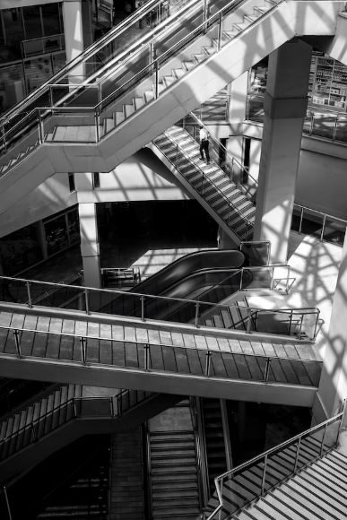 Black and white image of a person walking on interwoven staircases and escalators in a multi-level structure, with geometric shadows.