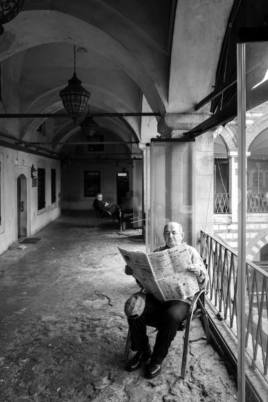 An elderly man sits in a chair under an archway, reading a newspaper. Another person is seated in the background. The setting has arched ceilings and an open window with a metal railing.