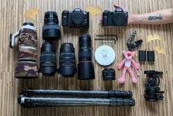 A collection of camera gear including two cameras, various lenses, a lens hood, a pink plush toy, a photographer's arm, tripods, and camera accessories on a wooden floor.