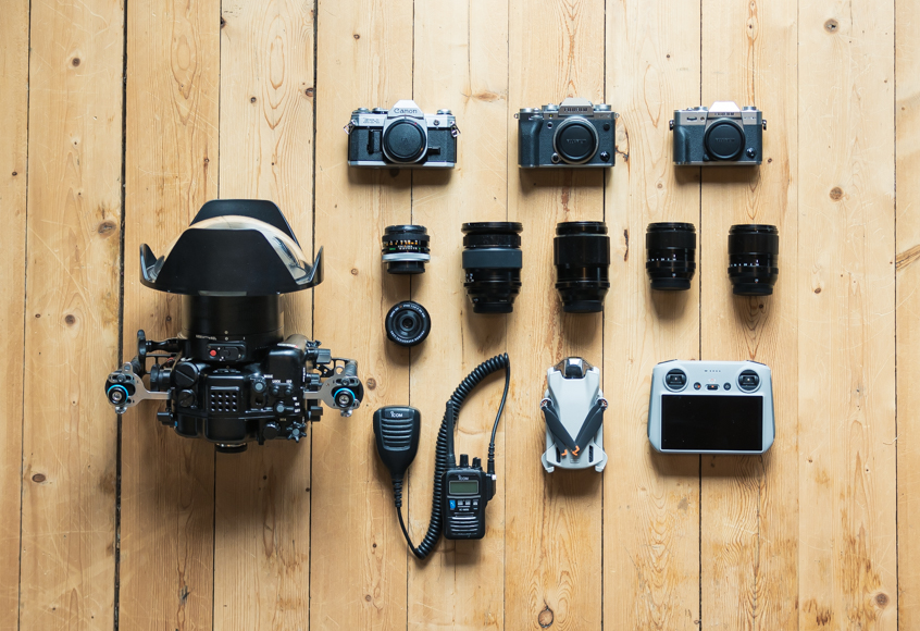 An overhead view of photography equipment arranged on a wooden floor, including cameras, lenses, a drone, a controller, and communication devices.