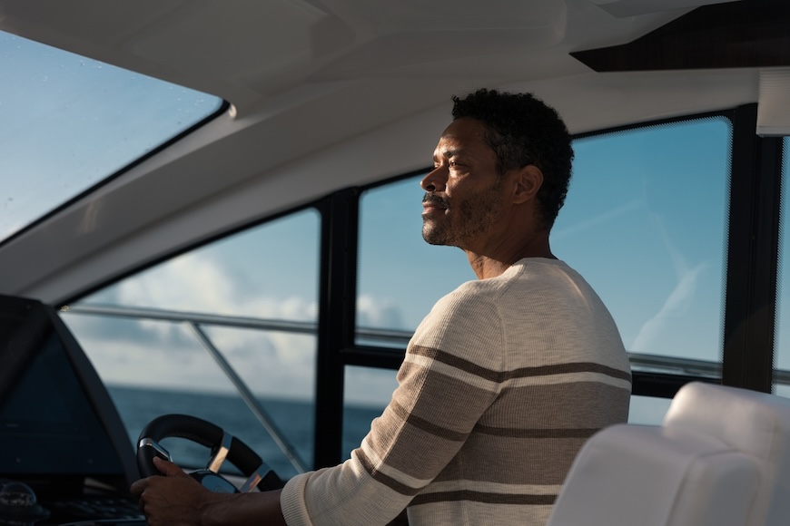 A man steers a boat, looking out to the sea. He is wearing a striped sweater.