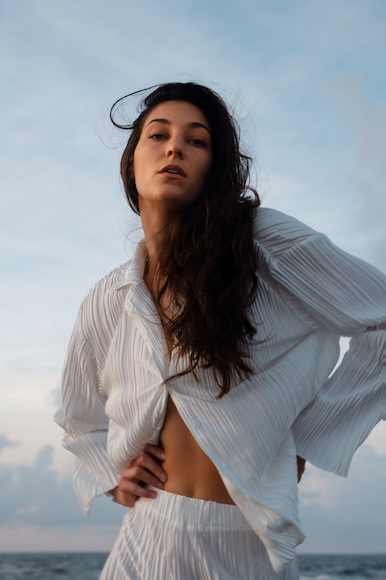 A person with long hair wearing a white outfit stands outdoors against a cloudy sky.