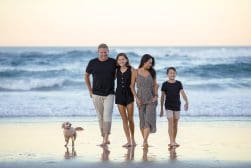 A family of four walks with a dog along a beach, smiling and holding hands. The ocean waves are in the background.