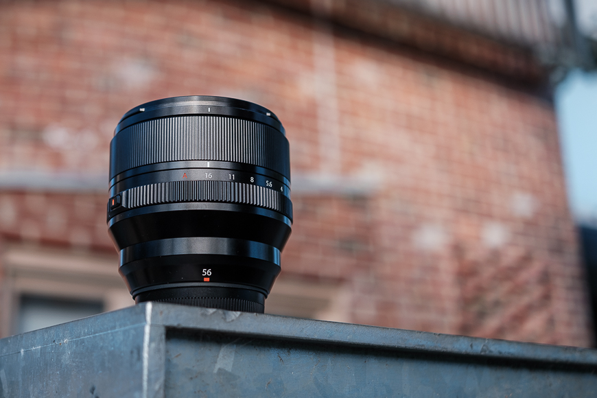 A 56mm camera lens resting on a metal railing, with a blurred brick building in the background.
