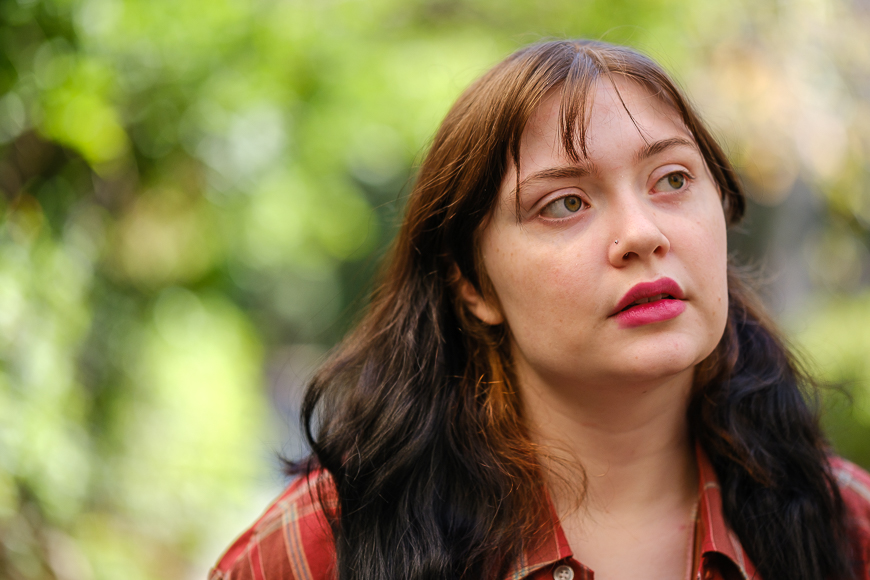 Young woman in a plaid shirt outdoors, looking away thoughtfully with a background of blurred green foliage.