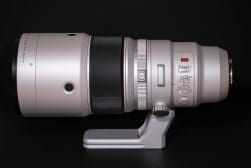 A large silver camera lens with a black focus ring and various control switches is mounted on a tripod foot against a dark background.