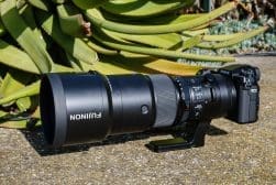 A Fujifilm camera with a large telephoto lens is mounted on a tripod support and positioned on a concrete surface next to green, spiky leaves.