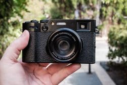 Hand holding a black rangefinder-style camera with a prime lens outdoors.