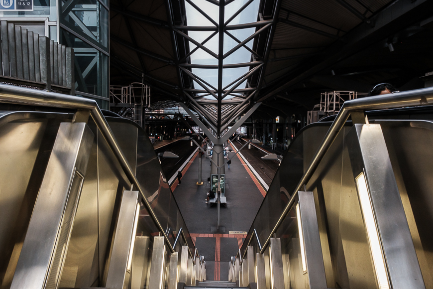 A view from the top of an escalator leads down to a modern train platform with multiple tracks. The station features metal and glass architecture.