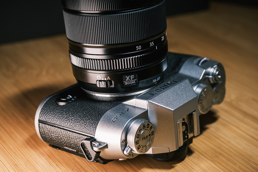 Close-up of a Fujifilm X-E4 camera with a Fujinon XF 16-80mm lens, lying on a wooden surface.