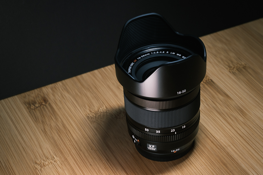 A black Fujinon XF 16-50mm camera lens with a hood, placed on a wooden surface with a dark background.