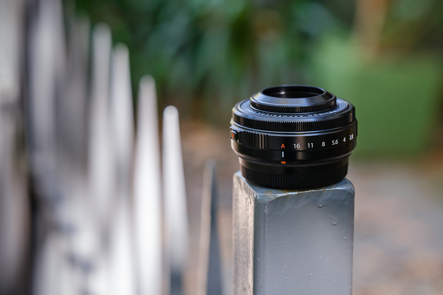 A camera lens rests on a fence post in a blurred natural background.