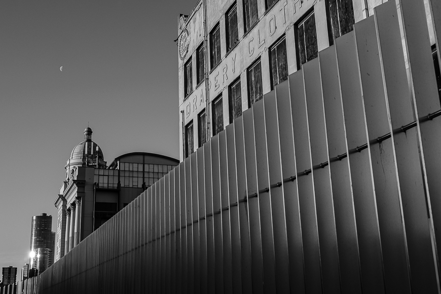 Black and white image of an urban scene featuring old buildings with a visible clock tower, modern metallic fencing in the foreground, and a crescent moon in the sky.