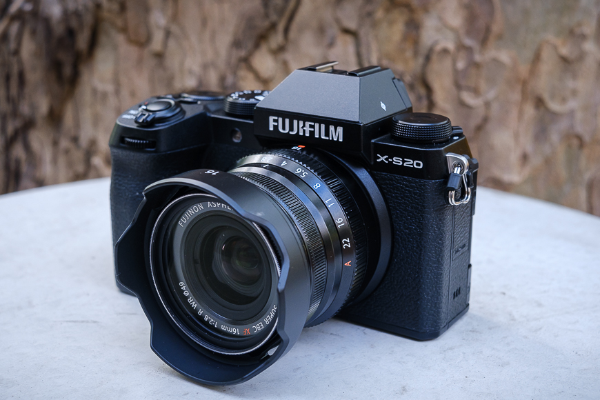 A fujifilm x-s20 mirrorless camera with a lens attached, displayed on a stone surface against a blurred bark background.