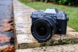 A Fujifilm X-T50 camera with a Fujinon aspherical lens placed on a stone surface, with fallen leaves nearby and green foliage in the background.