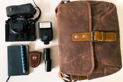 A brown camera bag, Nikon camera, external flash, notebook with pen, lens case, and lens cap arranged on a white surface.