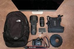A collection of camera gear including a Canon EOS 80D, three lenses, a camera harness, a backpack, a computer monitor, an external hard drive, a memory card, and a lens hood arranged on a surface.