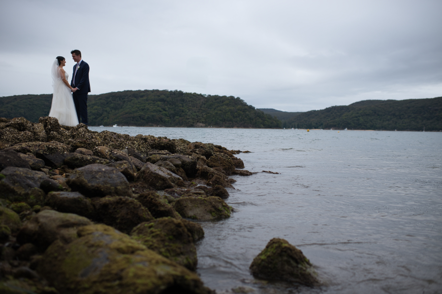 A couple in wedding attire stands on a rocky shoreline, holding hands, with a calm body of water and a hilly landscape in the background.