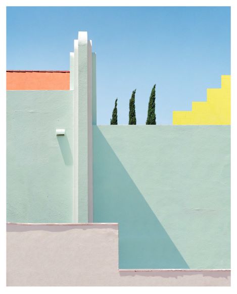 Abstract composition of colorful geometric building shapes with pastel green, yellow, and peach sections. Three tall trees are visible in the background against a clear blue sky.