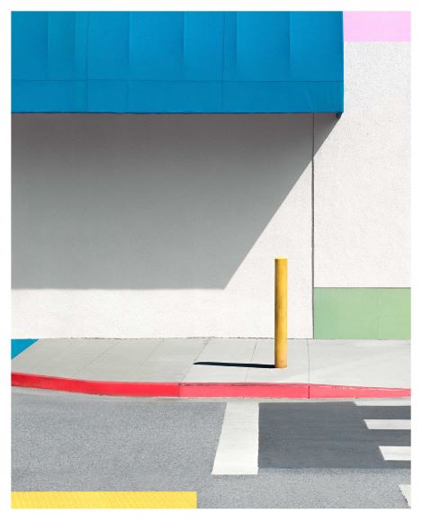 A minimalist urban scene with a yellow post casting a shadow on a sidewalk bordered by red and white lines, beneath a blue awning.