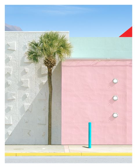 A minimalist urban scene with a palm tree, textured white wall, pink wall with three round fixtures, and a blue post against a clear sky.