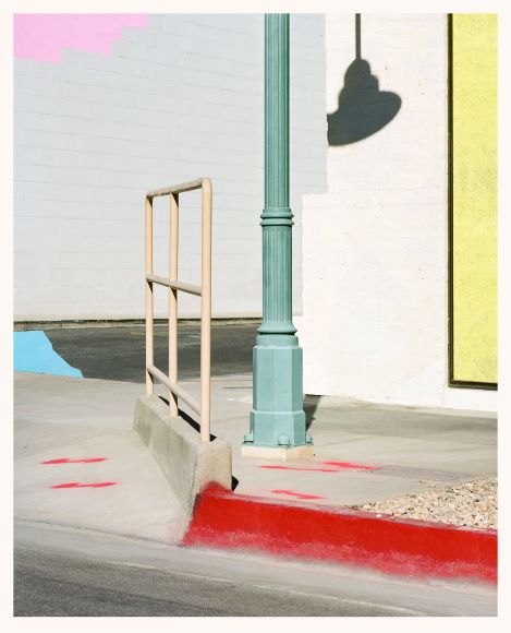 A teal streetlamp casts a shadow on a pale wall. A tan railing is next to a red painted curb with some red spots on the pavement. The scene is minimal and brightly lit.