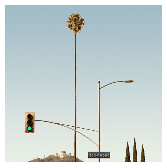 Tall palm tree next to a streetlight and traffic light at green, with a sign below reading "Hollywood." Hills with observatories visible in the background under a clear sky.
