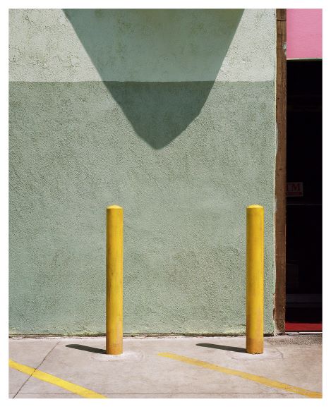 Two yellow bollards in front of a green wall with a shadow and a partial view of a door on the right.