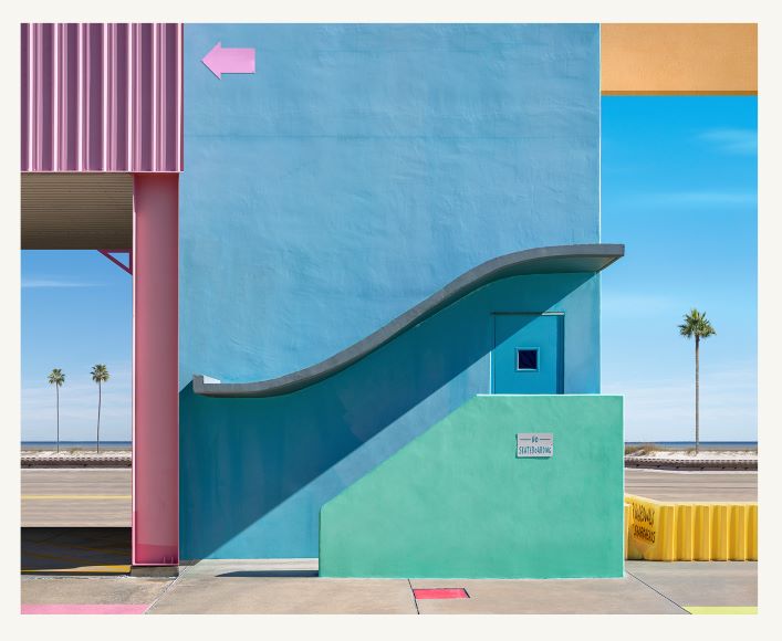 A colorful, geometric building features a wavy blue wall, a small window, and various pastel colors. Palm trees and a road appear in the background under a clear blue sky.