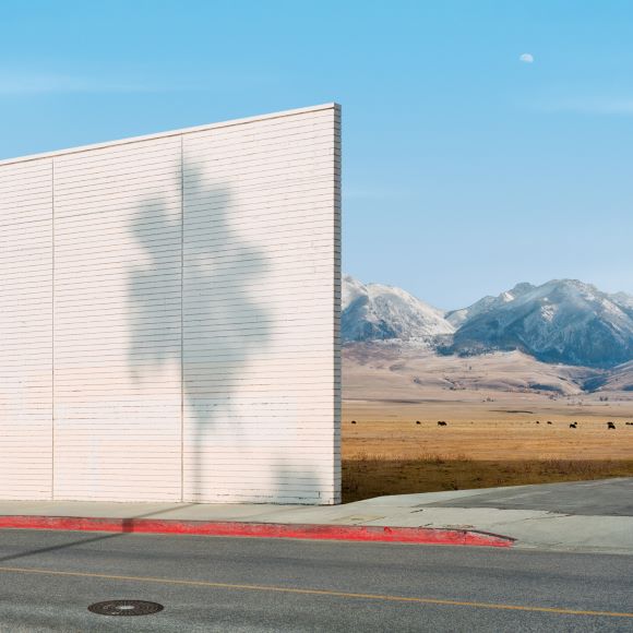 A white wall casts a shadow of a tree on a roadside curb. Behind it, a vast field stretches towards distant snow-capped mountains under a clear blue sky.