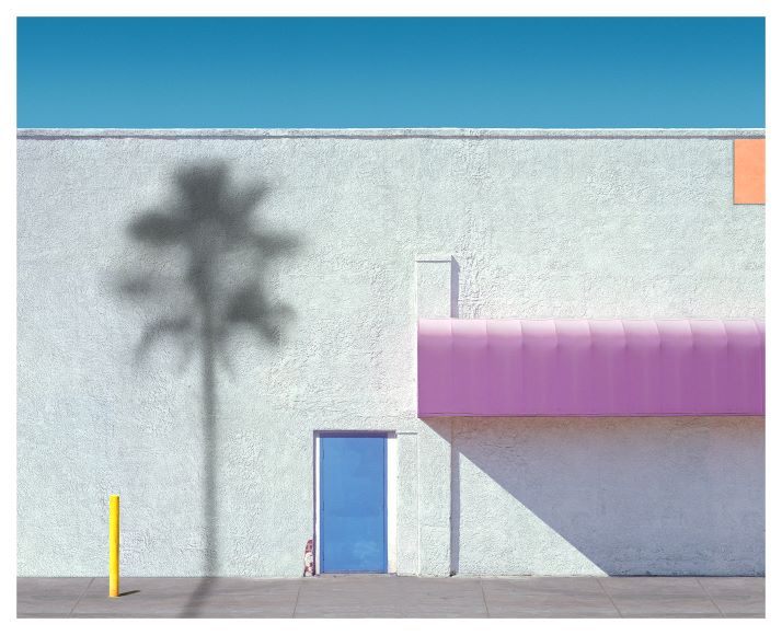 A minimalist scene with a white wall, small blue door, pink awning, and shadow of a palm tree. A single yellow post stands on the left, set against a clear blue sky.