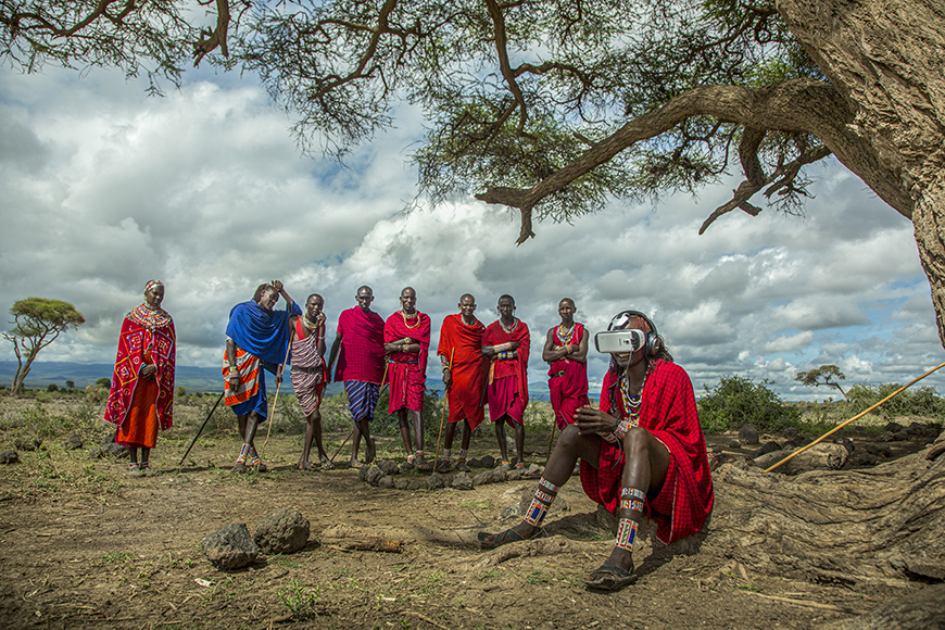 A group of people in colorful attire stand under a tree in a rural setting, with one person sitting and using a virtual reality headset.