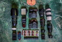 Camera equipment arranged on a textured surface, including two camera bodies, multiple lenses, flashes, batteries, memory cards, and a light panel. Bare feet visible at the top of the image.