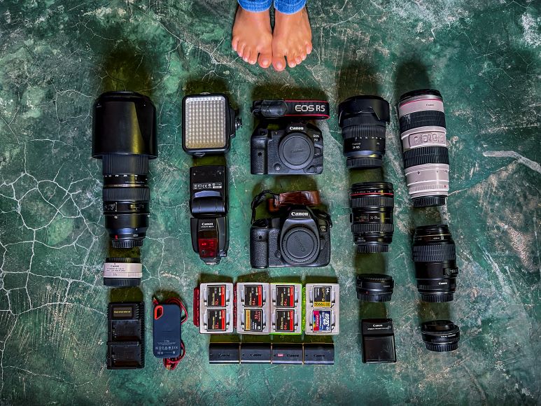 Camera equipment arranged on a textured surface, including two camera bodies, multiple lenses, flashes, batteries, memory cards, and a light panel. Bare feet visible at the top of the image.