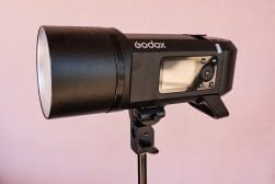 A black Godox studio flash unit with a digital display, mounted on a light stand, set against a light pink background.