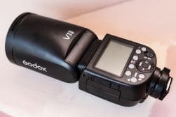 A Godox V1 flash unit, featuring a round head, LCD screen, control buttons, and a mount for attaching to a camera.