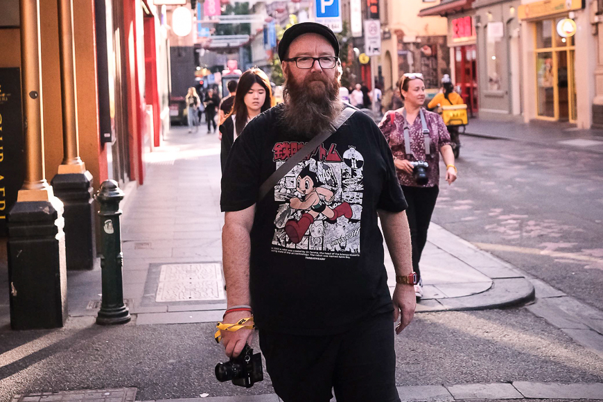 A bearded man wearing glasses and a graphic t-shirt walks on a bustling street holding a camera.