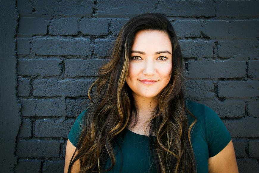 Woman with long hair wearing a green top standing against a dark brick wall.