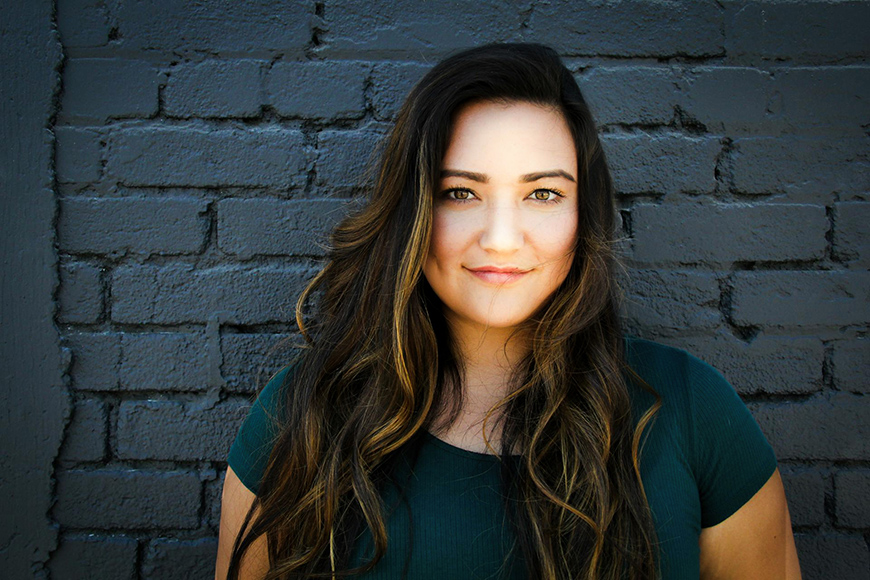 A woman with long brown hair and a green shirt stands in front of a gray brick wall, looking at the camera.