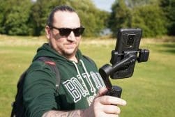 A person wearing sunglasses holds a smartphone mounted on a stabilizer in an outdoor park area.
