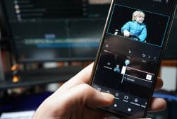 Hand edits a video featuring a toddler on a smartphone screen, with video editing software visible in the background on a computer monitor.