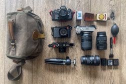 Camera equipment laid out on a wooden floor, including three cameras, three lenses, a tripod, a camera bag, film rolls, lens cleaning tools, and two battery chargers.