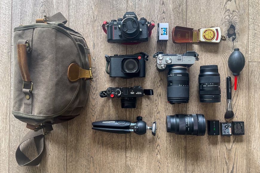 Camera equipment laid out on a wooden floor, including three cameras, three lenses, a tripod, a camera bag, film rolls, lens cleaning tools, and two battery chargers.